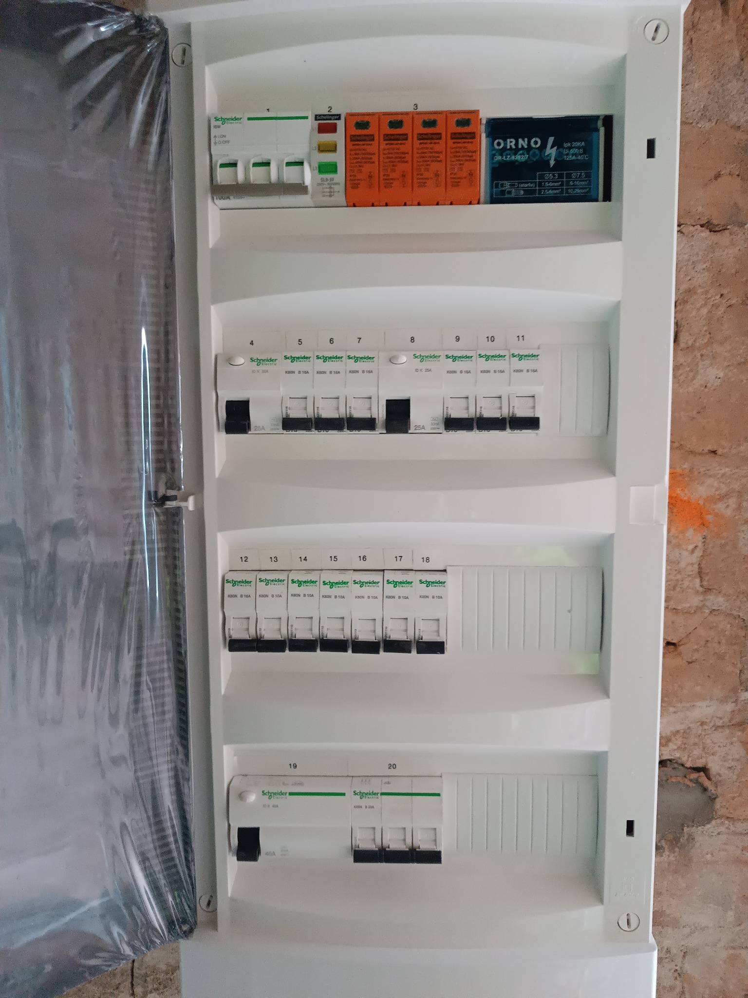 Otwarta skrzynka rozdzielcza z widocznymi bezpiecznikami, ogranicznikiem przepięć marki ORNO i zabezpieczeniami różnicowoprądowymi Schneider Electric, częściowo osłonięta folią ochronną...