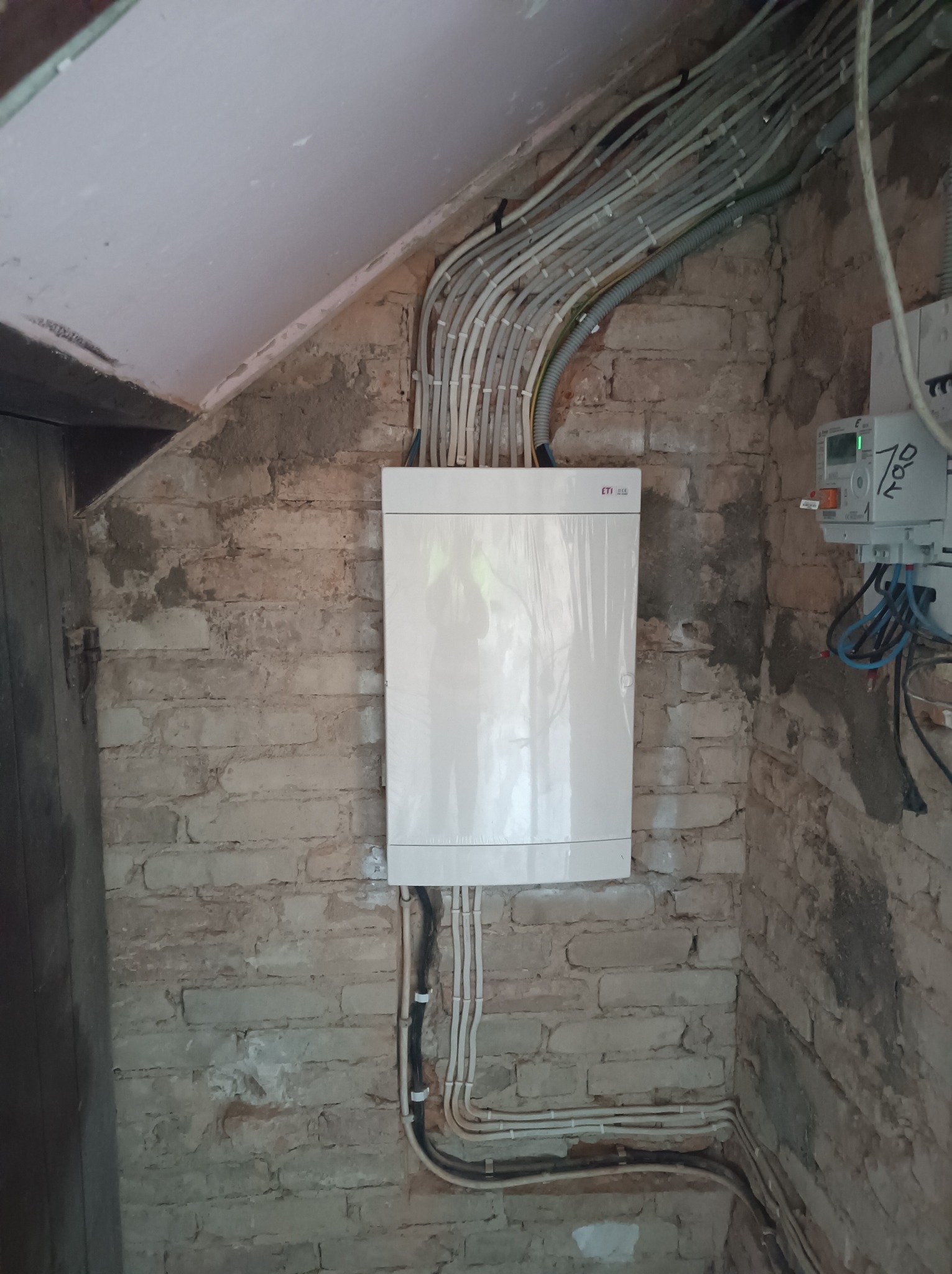 Biała skrzynka rozdzielcza ETI zamontowana na starej, ceglanej ścianie, otoczona wiązkami kabli elektrycznych w peszlach, z widocznym licznikiem energii elektrycznej po prawej stronie.