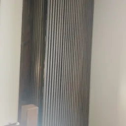 Panel ścienny z pionowych listew w odcieniach szarości, zamontowany w rogu pokoju, przylegający do jasnej ściany, widoczny fragment drewnianej konstrukcji.