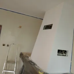 Świeżo pomalowany na biało komin z otworami na instalacje elektryczne, zabezpieczony folią, z widoczną drabiną i taśmą malarską na ścianach.