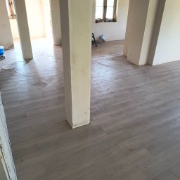 Leszek Mejer firma ogólnobudowlana - Ułożona podłoga z płytek imitujących drewno w jasnym odcieniu szarości, widoczne surowe ściany i filary w trakcie prac wykończeniowych, okno w tle.