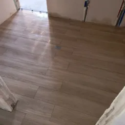 Ułożona podłoga z płytek imitujących drewno w łazience, widoczny odpływ liniowy i nieukończone obramowanie drzwi.