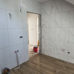 Leszek Mejer firma ogólnobudowlana - Łazienka w trakcie wykańczania: białe płytki kwadratowe na ścianach, imitujące marmur, drewnopodobne panele na podłodze, widoczne puszki elektryczne i wystające rury, w tle czerwone wiadro...