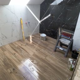 Leszek Mejer firma ogólnobudowlana - Łazienka w trakcie prac: ułożona podłoga imitująca drewno, marmurowe płytki na ścianach (białe i czarne), poziomica Stanley oparta o ścianę, ślady butów na podłodze, drabinka, rozdzielacz...