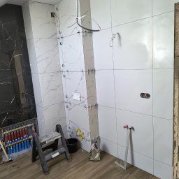 Leszek Mejer firma ogólnobudowlana - Łazienka w trakcie remontu: białe i czarne płytki imitujące marmur na ścianach, widoczne narzędzia glazurnicze, drabinka, instalacja grzewcza podłogowa i niezakończone podłączenia wodne.