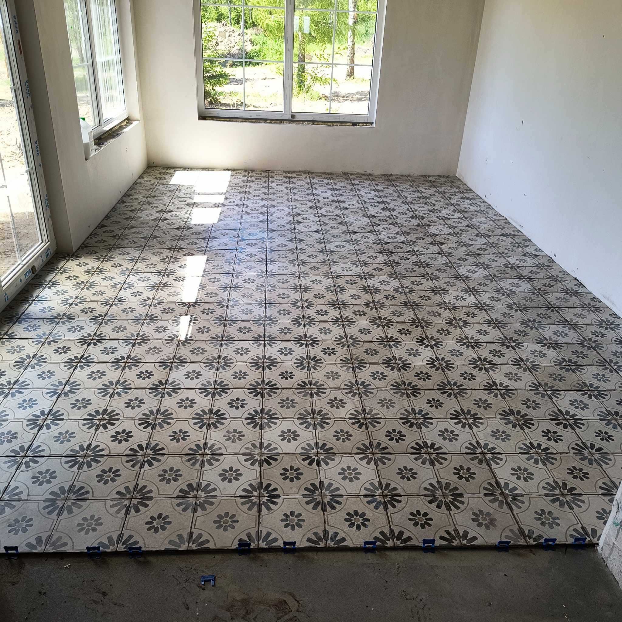 Układanie płytek podłogowych z kwiatowym wzorem w jasnym pomieszczeniu, widoczne niebieskie klipsy poziomujące, ślady światła słonecznego padające przez okno.