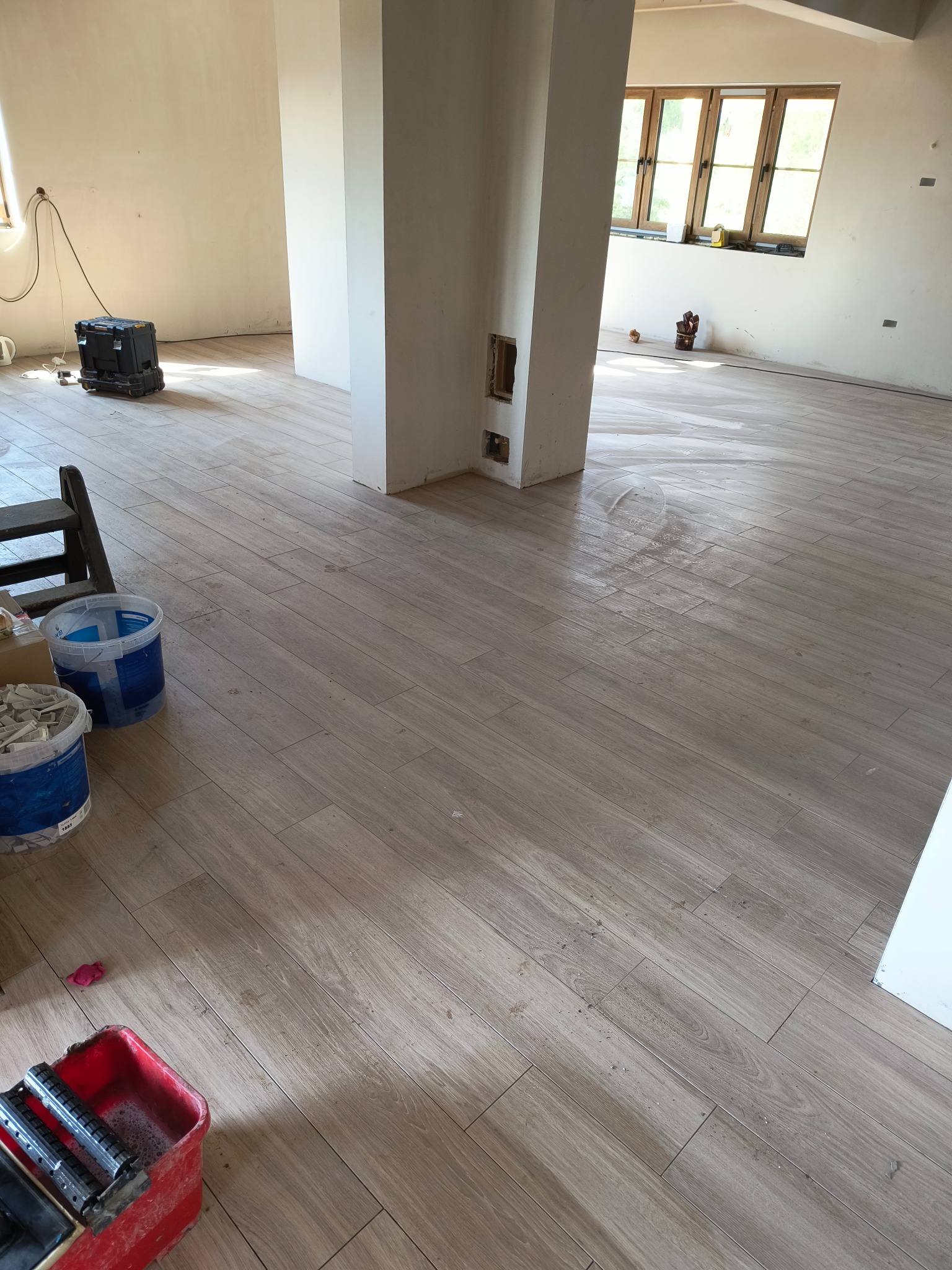 Układanie podłogi z płytek imitujących drewno w jasnym, przestronnym wnętrzu, widoczne narzędzia glazurnicze i ślady po czyszczeniu płytek.