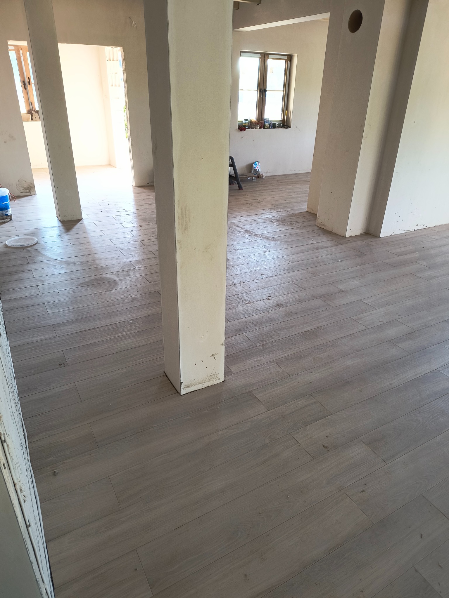 Ułożona podłoga z płytek imitujących drewno w jasnym odcieniu szarości, widoczne surowe ściany i filary w trakcie prac wykończeniowych, okno w tle.