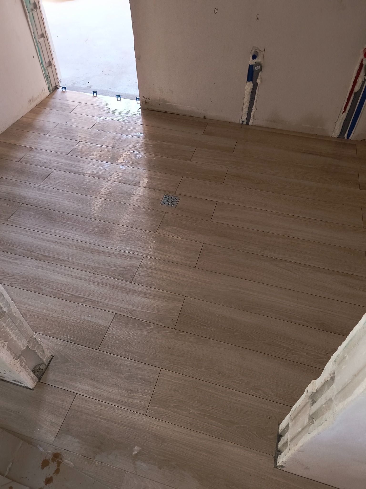 Ułożona podłoga z płytek imitujących drewno w łazience, widoczny odpływ liniowy i nieukończone obramowanie drzwi.