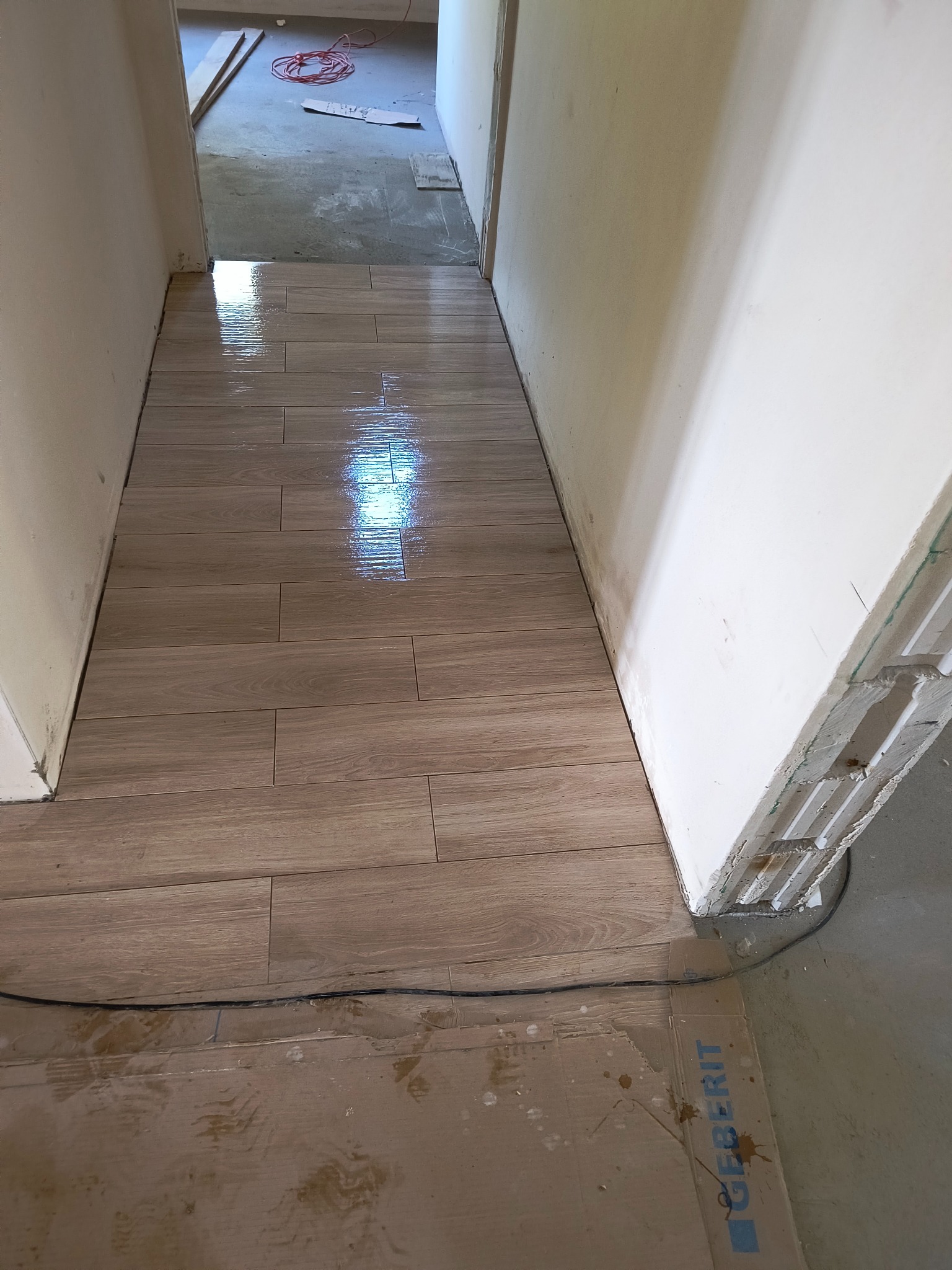 Ułożone podłużne płytki podłogowe imitujące drewno, świeżo po położeniu, z widocznymi refleksami wilgoci, w wąskim korytarzu w trakcie prac wykończeniowych, zabezpieczone kartonem z logo Geberit.