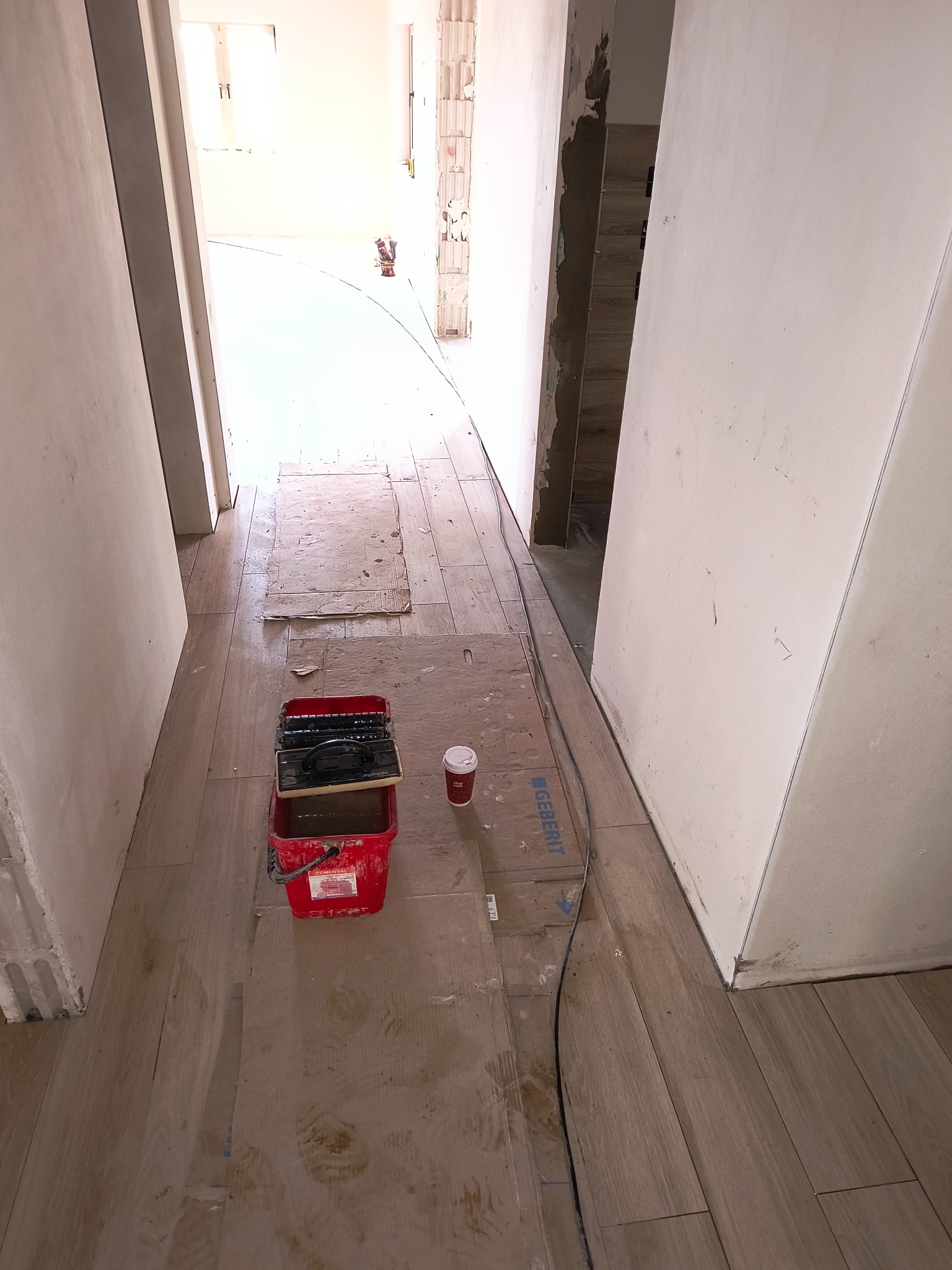 Podłoga w trakcie układania płytek, widoczny czerwony pojemnik z narzędziami, kubek po kawie i kabel Geberit