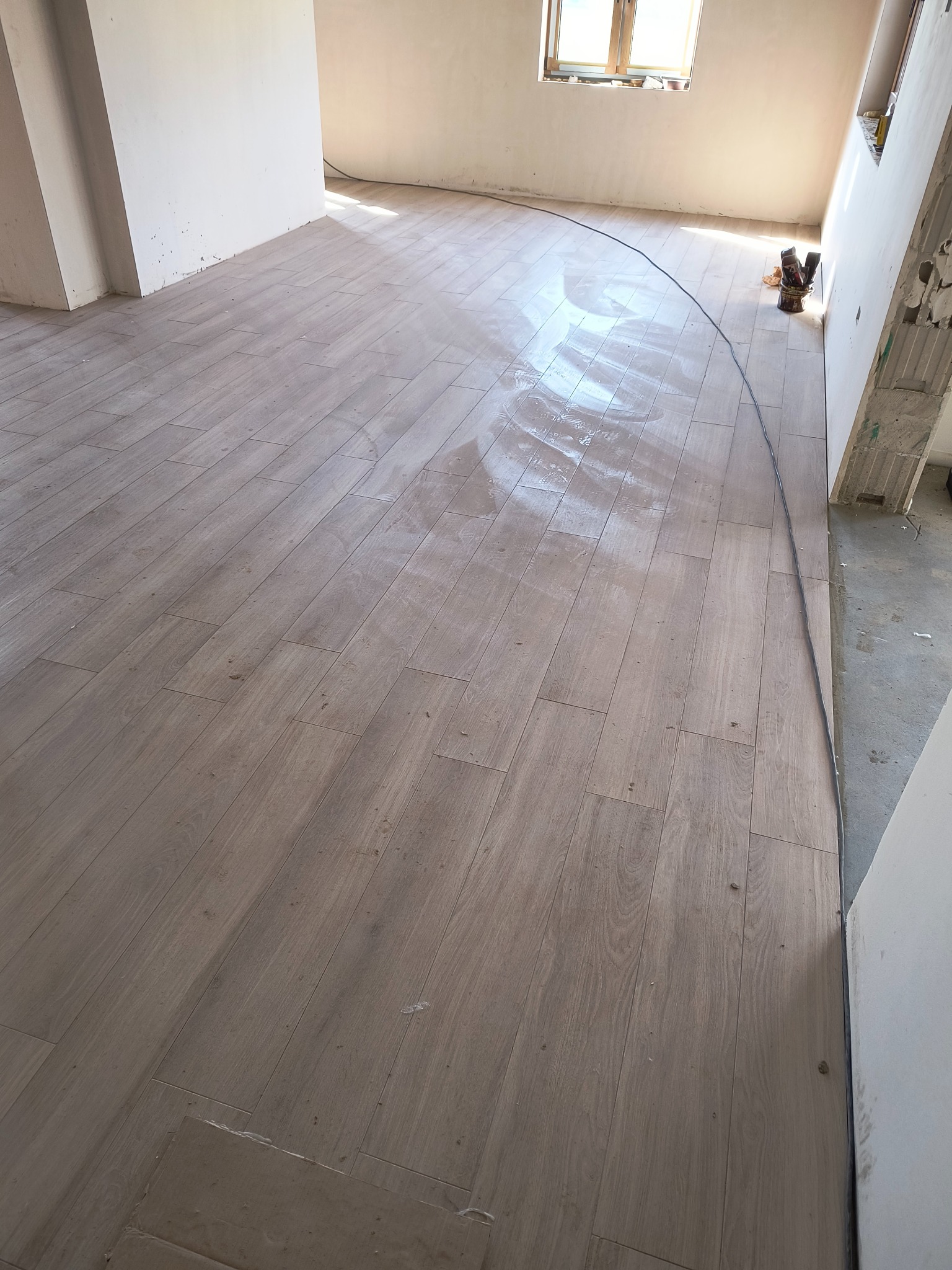 Ułożona podłoga z płytek imitujących drewno w jasnym odcieniu, widoczne niedoskonałości na powierzchni płytek, ściany w stanie surowym, okno w tle.