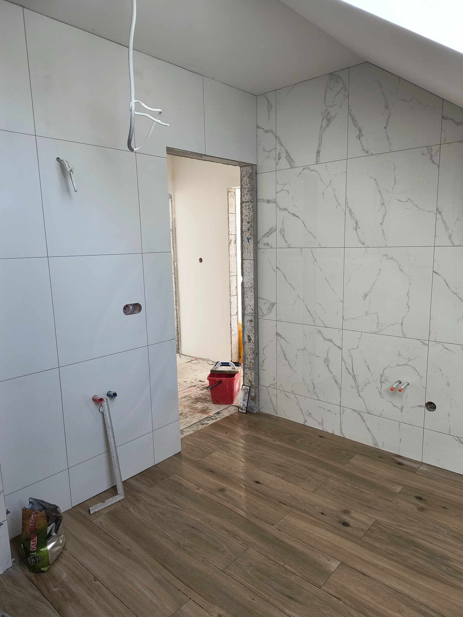 Łazienka w trakcie wykańczania: białe płytki kwadratowe na ścianach, imitujące marmur, drewnopodobne panele na podłodze, widoczne puszki elektryczne i wystające rury, w tle czerwone wiadro...