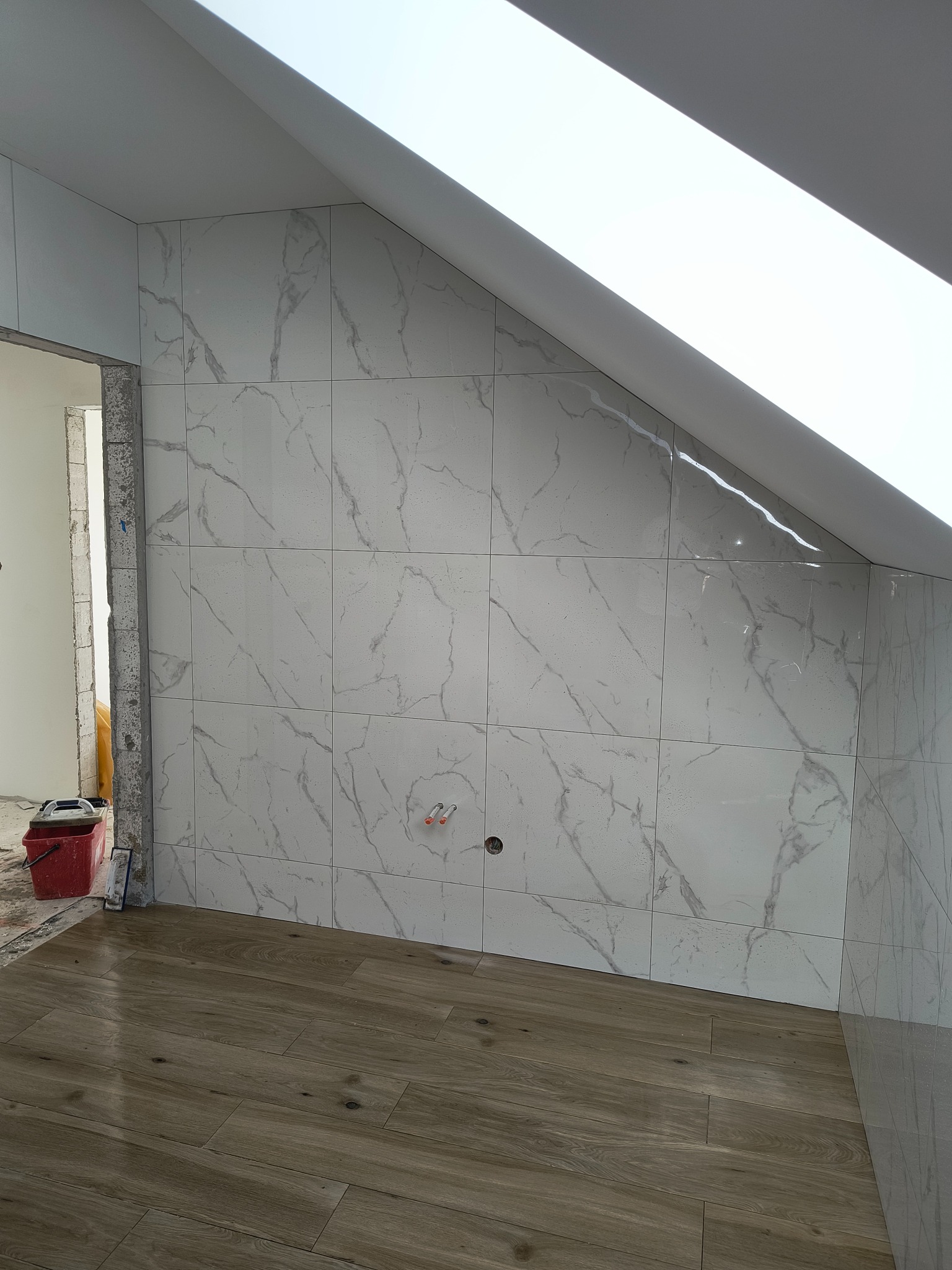 Ściana wyłożona białymi płytkami imitującymi marmur w łazience na poddaszu w Sierakowicach, widoczna drewnopodobna podłoga i fragment okna dachowego.