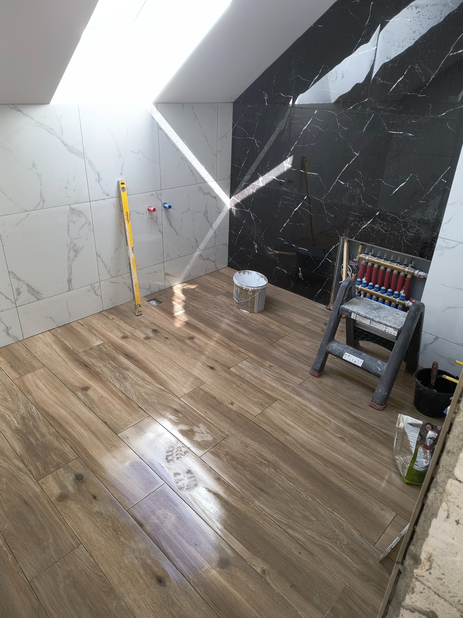 Łazienka w trakcie prac: ułożona podłoga imitująca drewno, marmurowe płytki na ścianach (białe i czarne), poziomica Stanley oparta o ścianę, ślady butów na podłodze, drabinka, rozdzielacz...