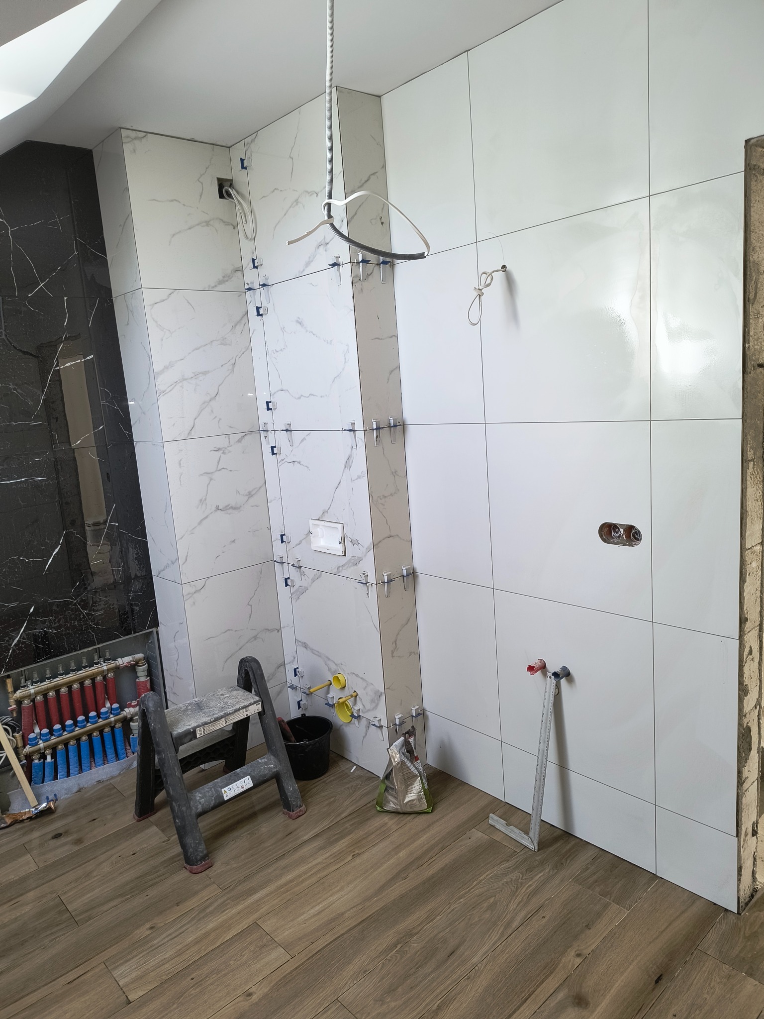 Łazienka w trakcie remontu: białe i czarne płytki imitujące marmur na ścianach, widoczne narzędzia glazurnicze, drabinka, instalacja grzewcza podłogowa i niezakończone podłączenia wodne.