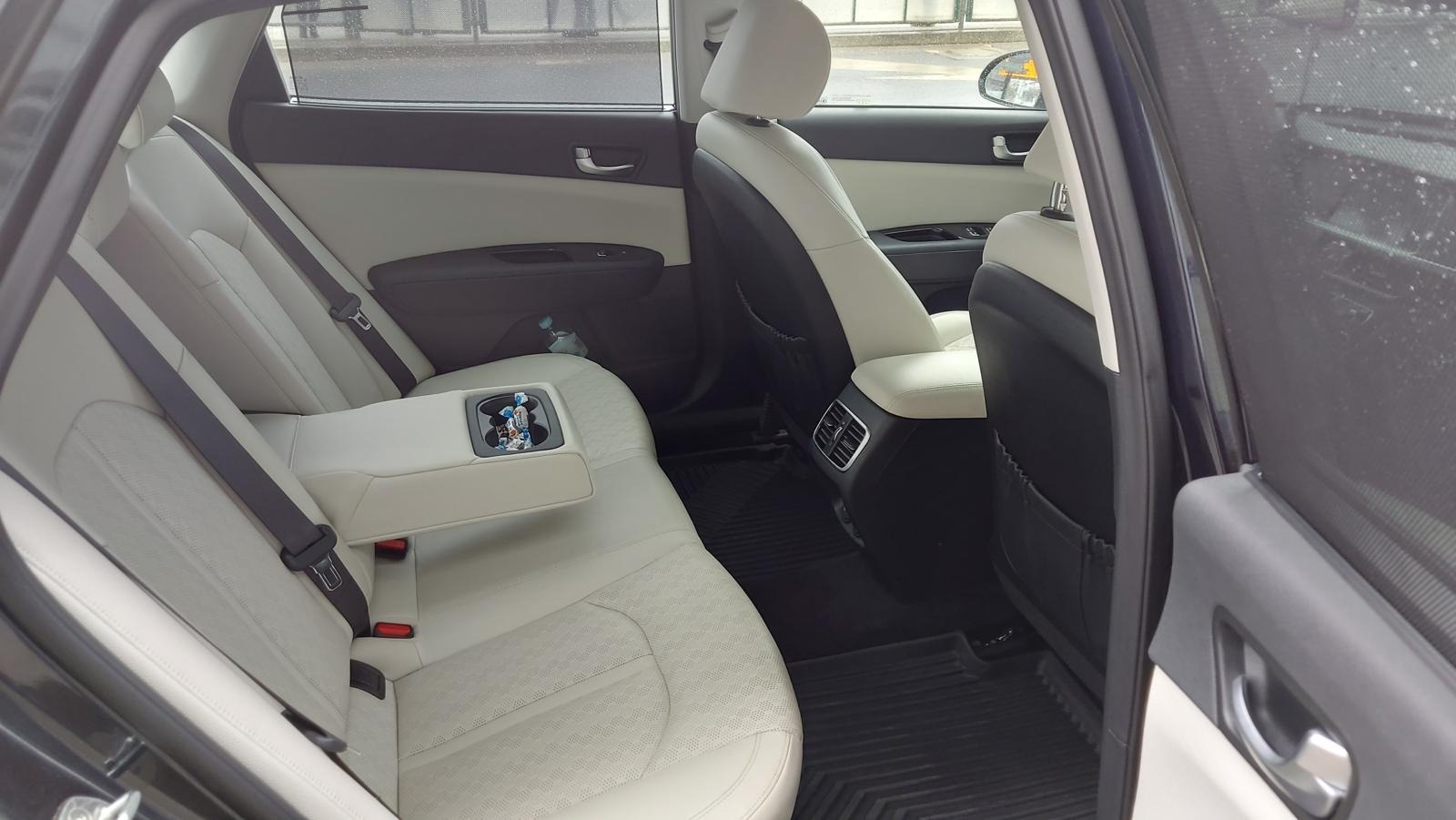 Wnętrze samochodu z jasną skórzaną tapicerką tylnych siedzeń, widoczne pasy bezpieczeństwa, podłokietnik z uchwytem na napoje zawierającym cukierki, oraz zasłonka przeciwsłoneczna w drzwiach.
