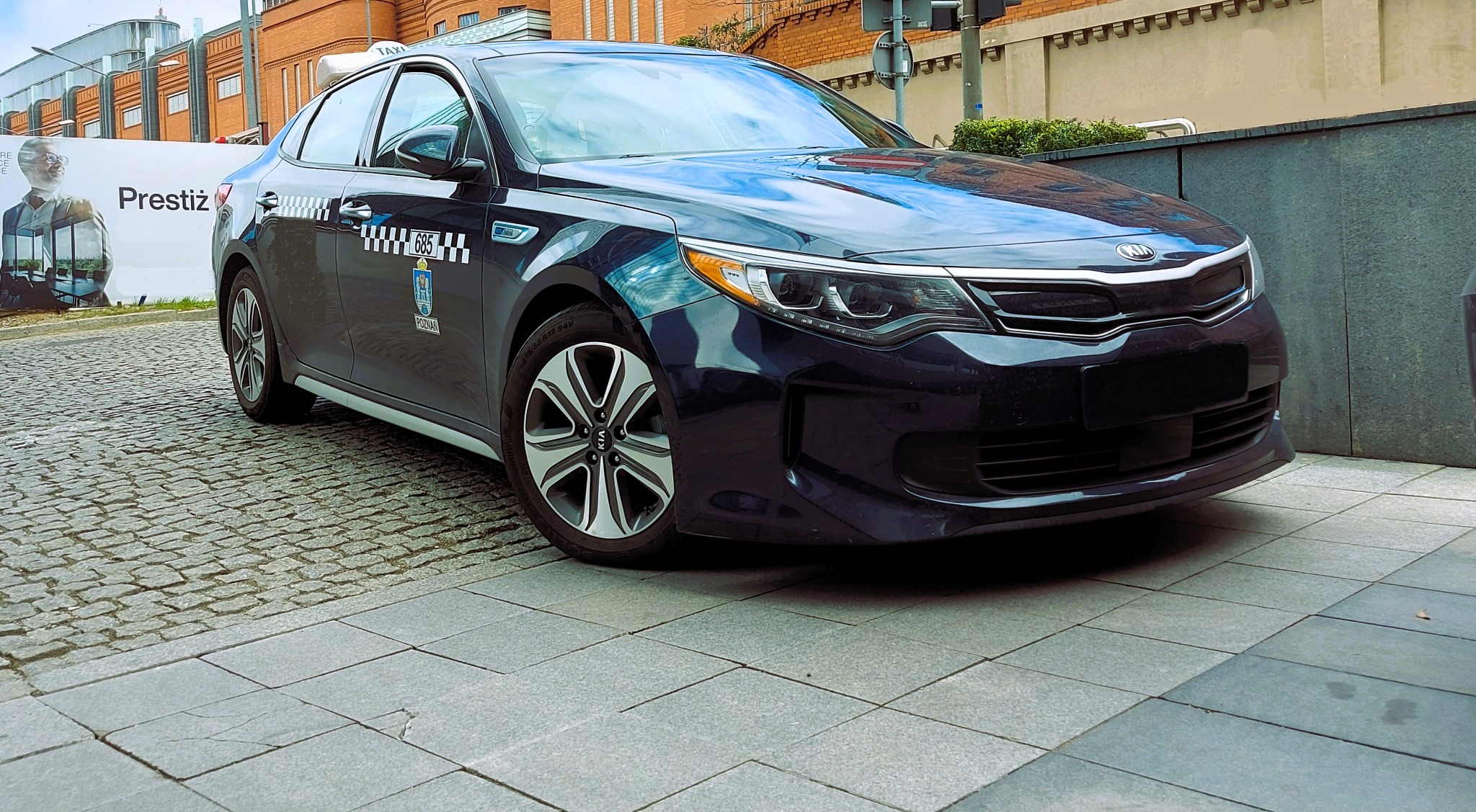 Ciemnogranatowy samochód osobowy Kia Optima z oznaczeniami taksówki, w tym herb Poznania, zaparkowany na chodniku z kostki brukowej i płyt.