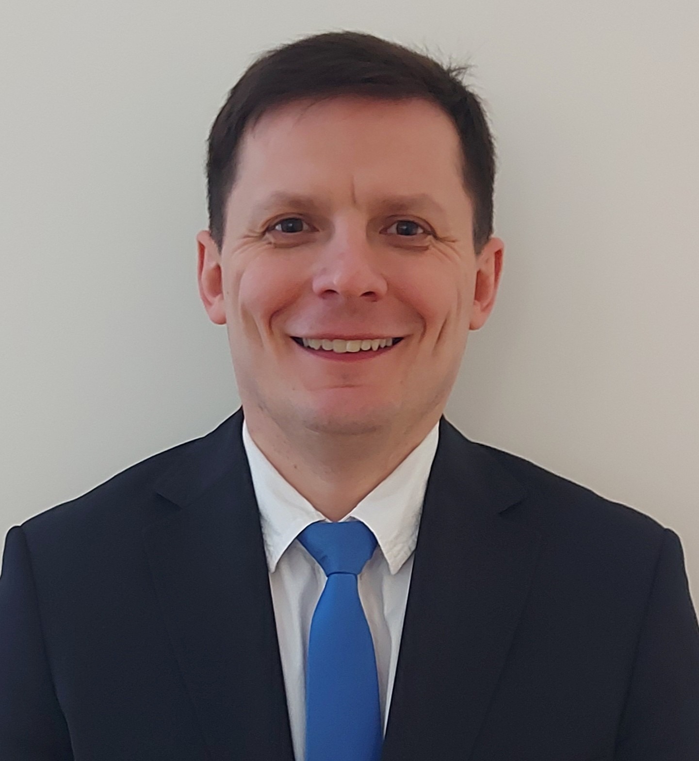 Uśmiechnięty mężczyzna w garniturze i niebieskim krawacie na jasnym tle.