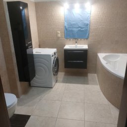 USŁUGI REMONTOWO BUDOWLANE ROBERT PIOTROWSKI - Jasna łazienka z beżowymi płytkami, pralką, umywalką z szafką i narożną wanną. Na ścianie roleta w kolorze niebieskim. Widok zza framugi drzwi.