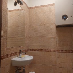 USŁUGI REMONTOWO BUDOWLANE ROBERT PIOTROWSKI - Beżowa łazienka z umywalką, lustrem i piecem gazowym Junkers. Ściany wyłożone mozaiką w odcieniach beżu i brązu. W koszyku pod umywalką rolki papieru toaletowego.