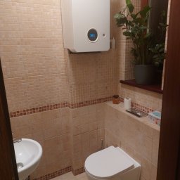 USŁUGI REMONTOWO BUDOWLANE ROBERT PIOTROWSKI - Beżowa łazienka z mozaiką, wiszącą toaletą i umywalką. Na ścianie piec gazowy Junkers. Uroku dodaje roślina doniczkowa na parapecie.