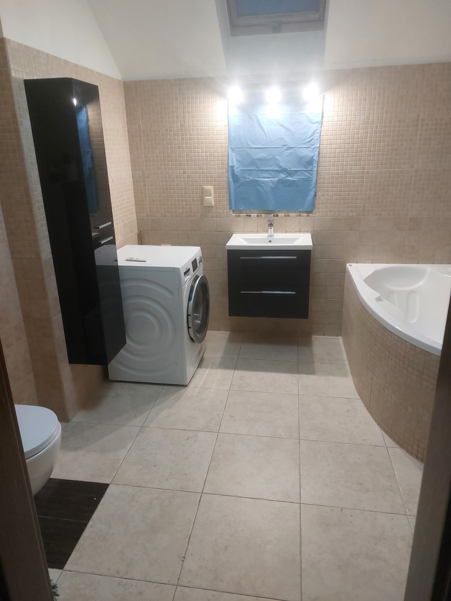 Jasna łazienka z beżowymi płytkami, pralką, umywalką z szafką i narożną wanną. Na ścianie roleta w kolorze niebieskim. Widok zza framugi drzwi.