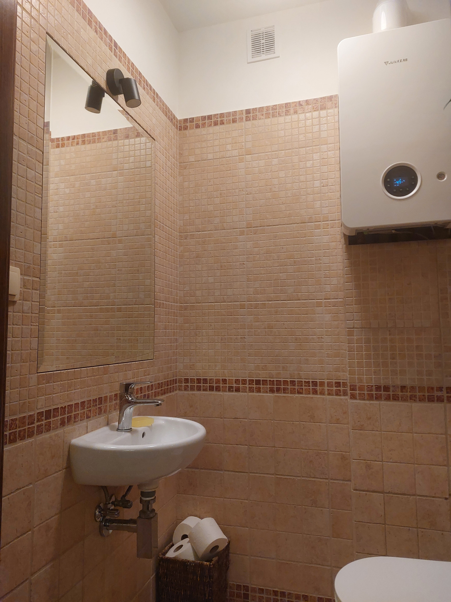 Beżowa łazienka z umywalką, lustrem i piecem gazowym Junkers. Ściany wyłożone mozaiką w odcieniach beżu i brązu. W koszyku pod umywalką rolki papieru toaletowego.
