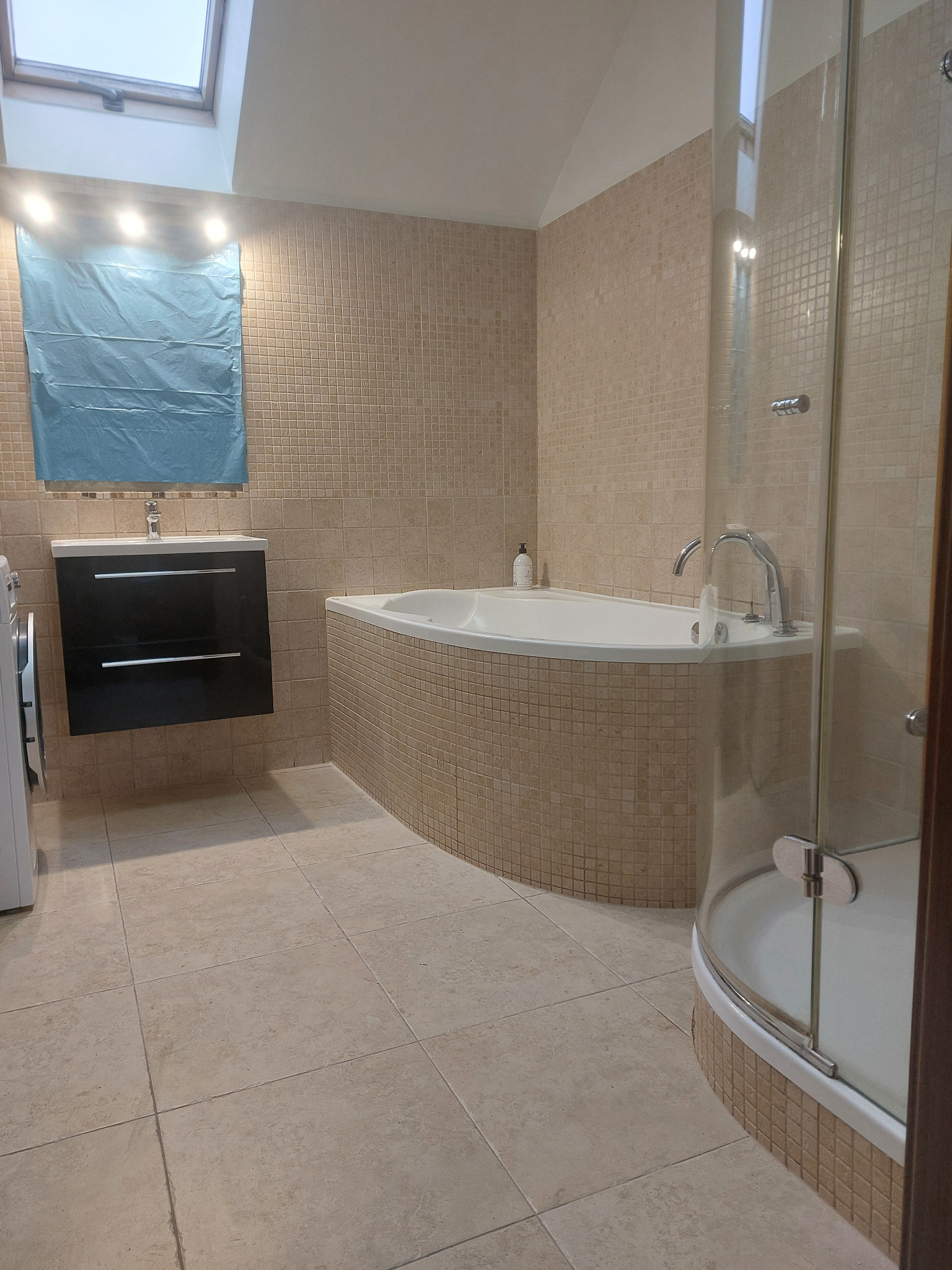 Jasna łazienka z mozaiką, kabiną prysznicową i wanną narożną. Umywalka z szafką w kolorze czarnym. Okno dachowe. Całość utrzymana w ciepłej tonacji beżu i brązu.