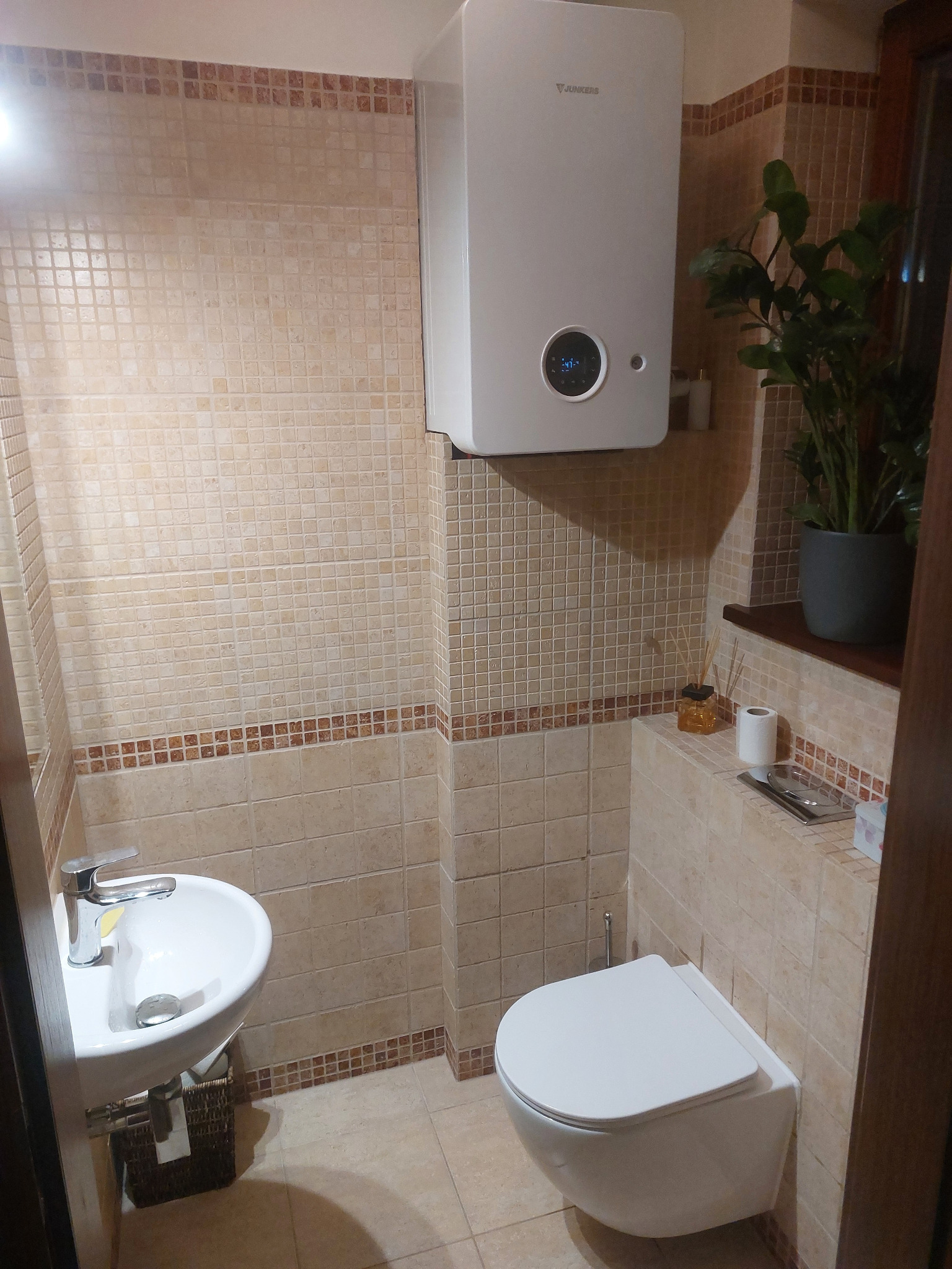 Beżowa łazienka z mozaiką: wisząca toaleta, umywalka nablatowa, piec gazowy Junkers, roślina doniczkowa. Ciepłe światło, przytulne wnętrze.