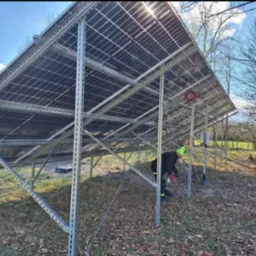 Montaż paneli fotowoltaicznych na konstrukcji stalowej, widok z dołu. Pracownik w zielonej czapce wykonuje prace montażowe. Słoneczny dzień, widoczne drzewa w tle.