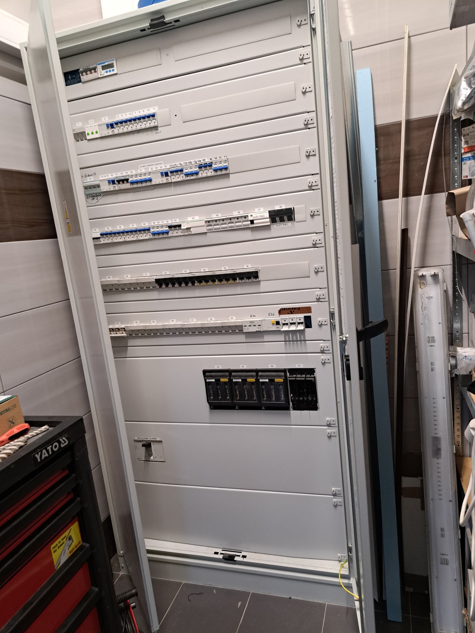 Rozdzielnica elektryczna z widocznymi bezpiecznikami, wyłącznikami i okablowaniem, częściowo otwarta, obok skrzynka narzędziowa YATO i regał z elementami instalacyjnymi.