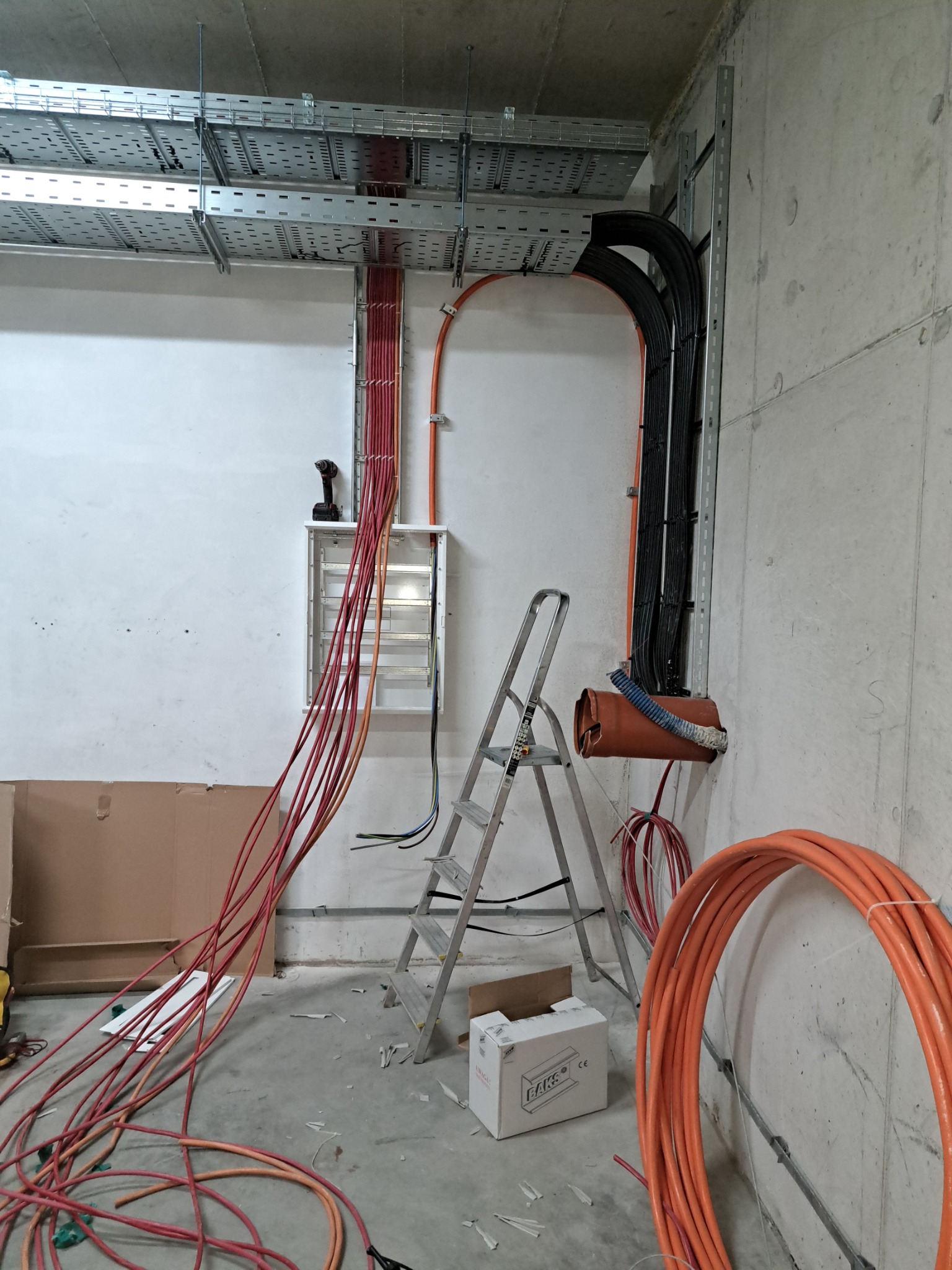 Montaż instalacji elektrycznej: widok na metalowe koryta kablowe pod sufitem, prowadzone kable w kolorach czerwonym i czarnym, skrzynka rozdzielcza z wiertarką na górze, drabina aluminiowa, zwój...