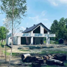 Surowy stan domu z betonu komórkowego w trakcie budowy, widoczna drabina oparta o ścianę, drewno na pierwszym planie, błękitne niebo w tle.