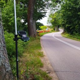 GEO-TARGET Usługi geodezyjne mgr inż. Arkadiusz Bartnicki - Pomiar geodezyjny przy drodze: tyczka z odbiornikiem GPS i kontrolerem, w tle widoczny geodeta w kamizelce pomiarowej i samochód.