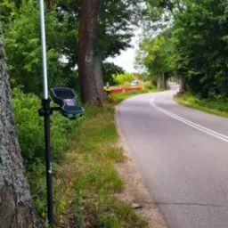 Pomiar geodezyjny przy drodze: tyczka z odbiornikiem GPS i kontrolerem, w tle widoczny geodeta w kamizelce pomiarowej i samochód.