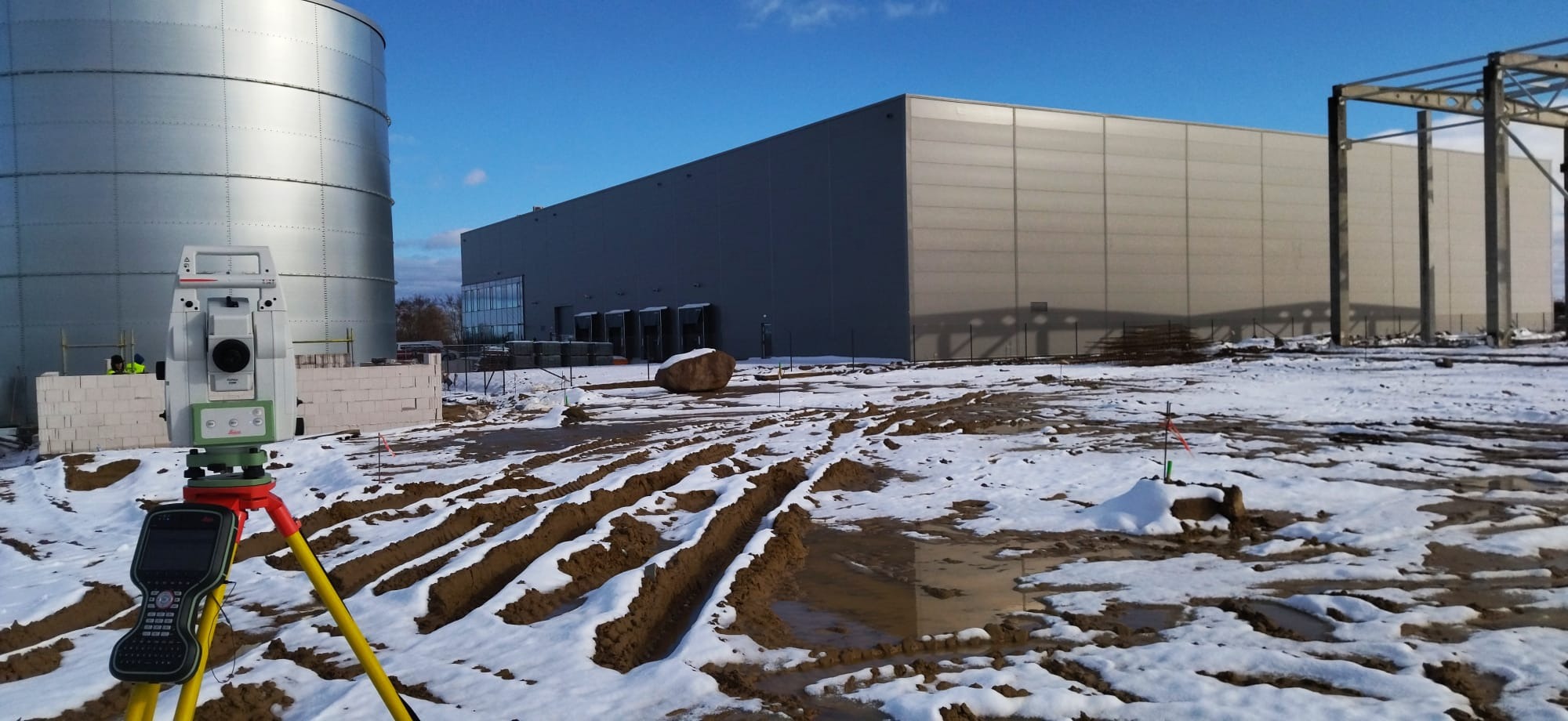 Tachimetr Leica na statywie, w tle budowa hali magazynowej i silos, teren częściowo pokryty śniegiem.