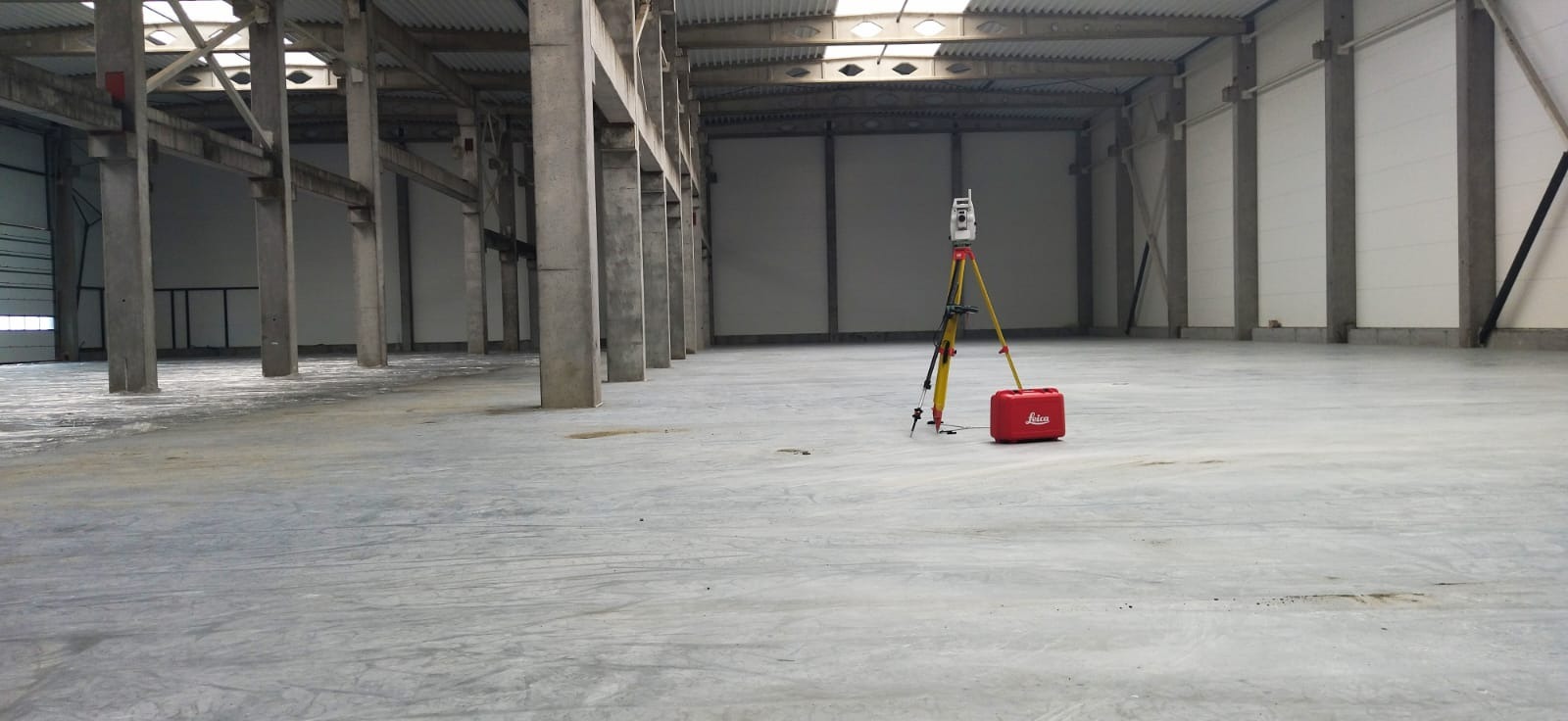 Wnętrze hali produkcyjnej z betonową posadzką, gdzie stoi żółty statyw z precyzyjnym instrumentem pomiarowym Leica i czerwona skrzynka na narzędzia.