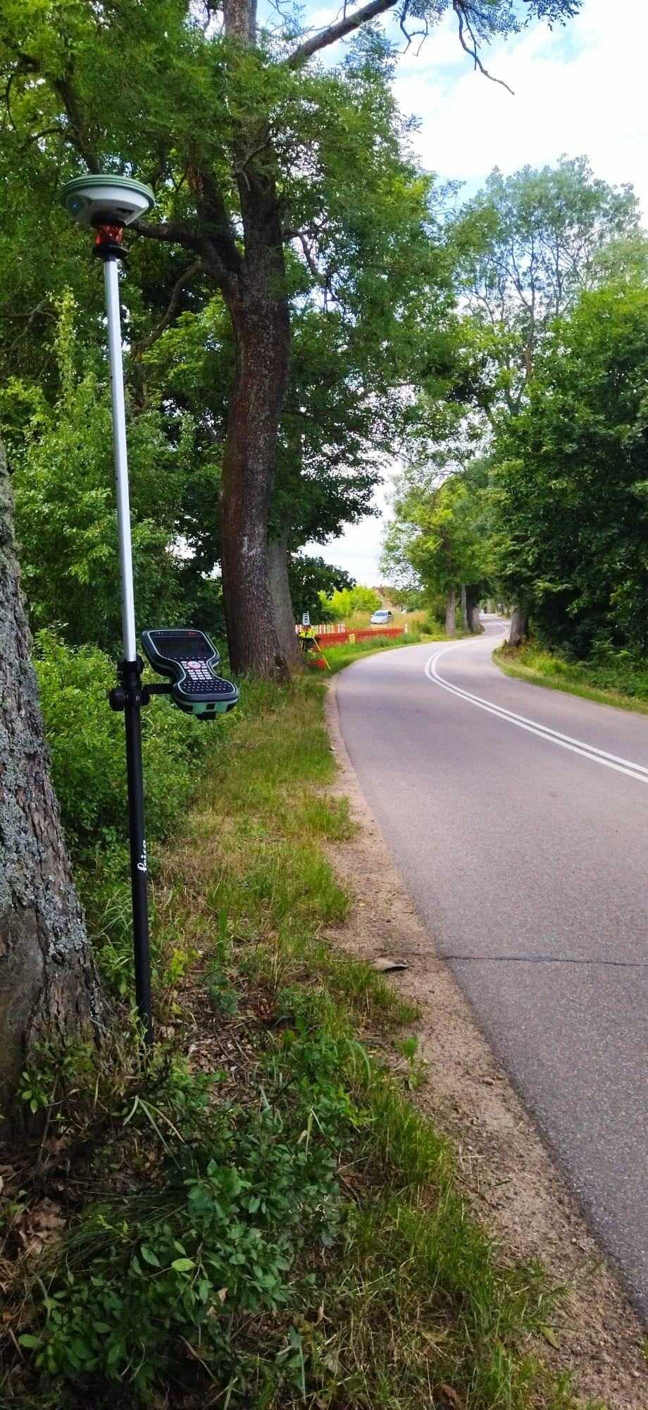 Pomiar geodezyjny przy drodze: tyczka z odbiornikiem GPS i kontrolerem, w tle widoczny geodeta w kamizelce pomiarowej i samochód.