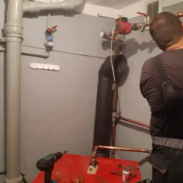 Instalator w trakcie montażu instalacji grzewczej, widoczny fragment kotła, rury miedziane, narzędzia i elementy instalacji, ściana pomalowana szarą farbą.
