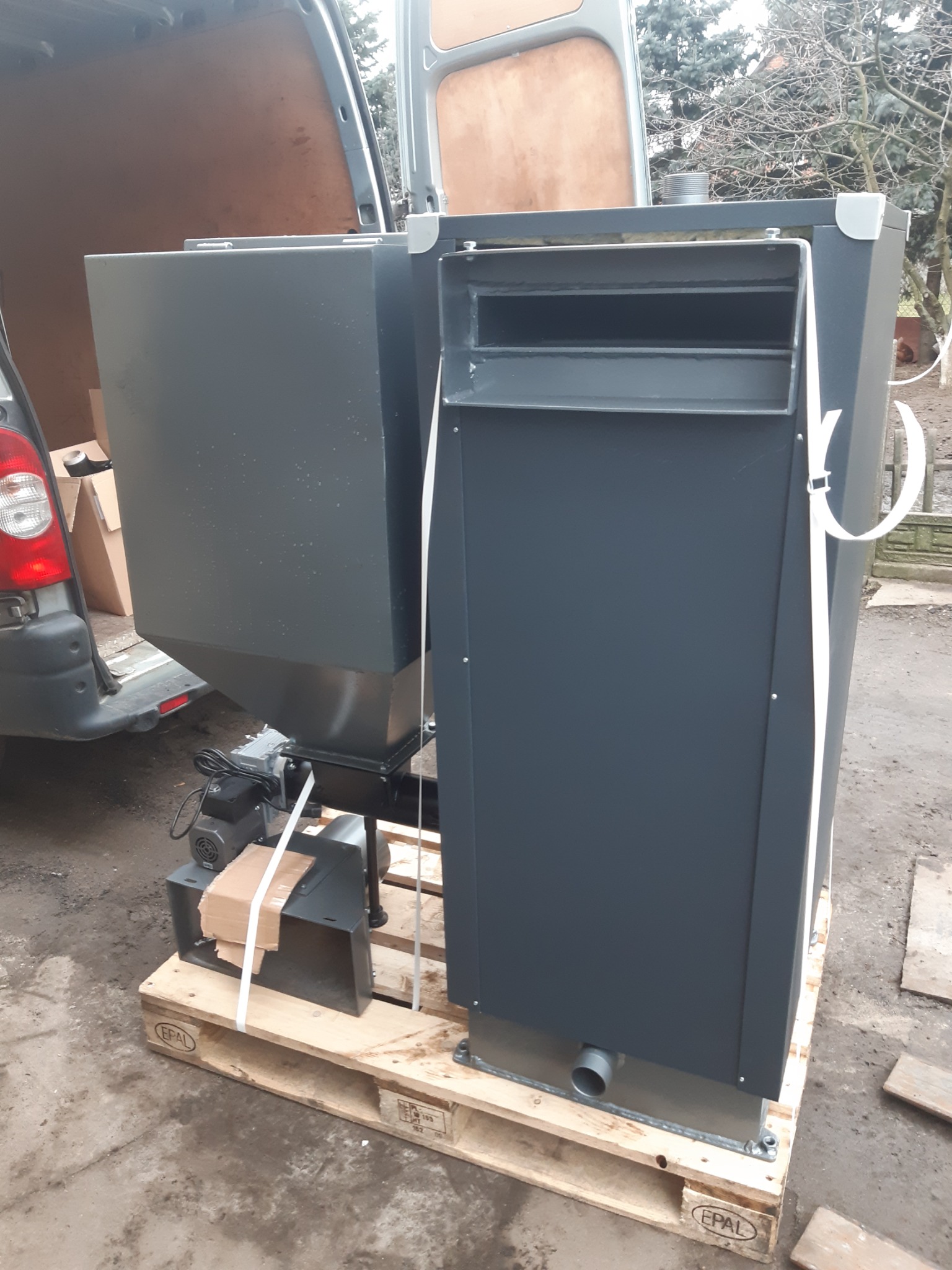 Nowy, szary piec na paliwo stałe z podajnikiem ślimakowym, umieszczony na palecie EPAL, wyładowywany z furgonetki na zewnątrz.