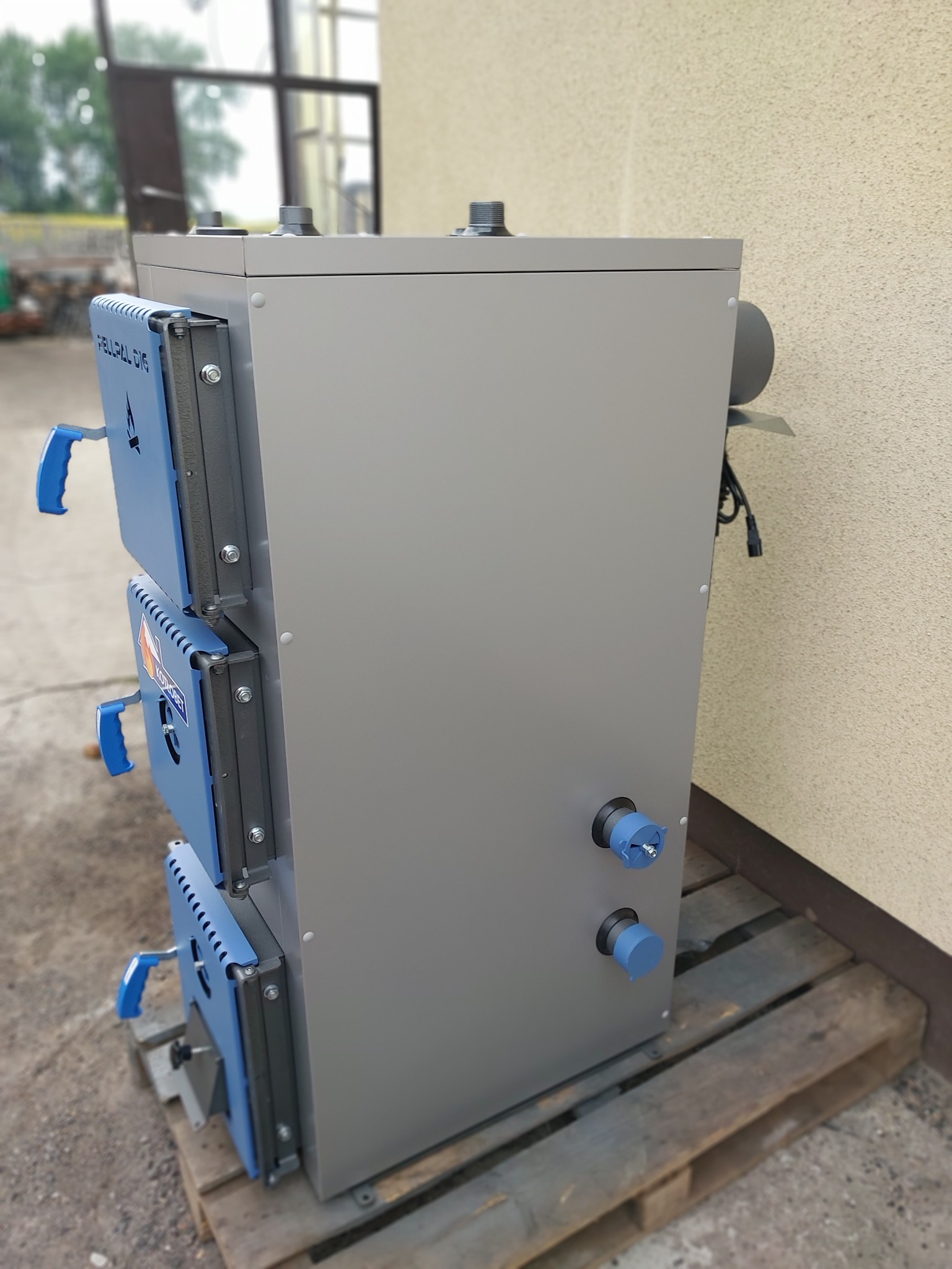 Nowoczesny piec na paliwo stałe, szary z niebieskimi detalami, ustawiony na palecie przy ścianie budynku.