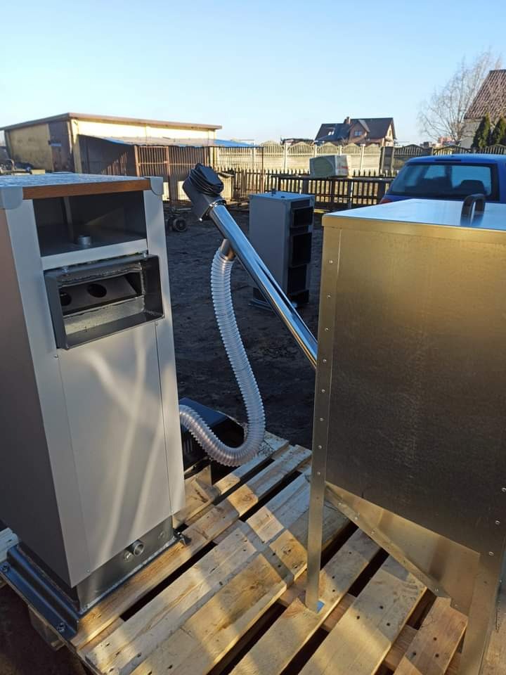 Dwa piece na paliwo stałe, połączone metalową rurą z przezroczystym wężem, ustawione na drewnianej palecie na zewnątrz, widoczne budynki gospodarcze i samochód w tle.