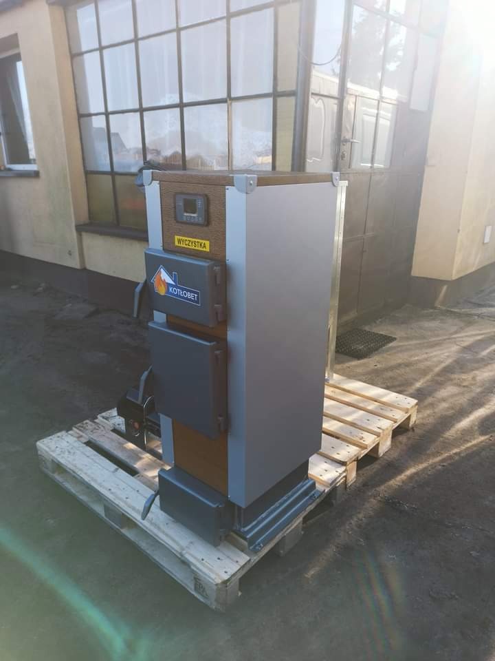 Szary piec na paliwo stałe marki Kotlobet, ustawiony na palecie przed budynkiem z dużym oknem, widoczna naklejka 'Wyczystka'.
