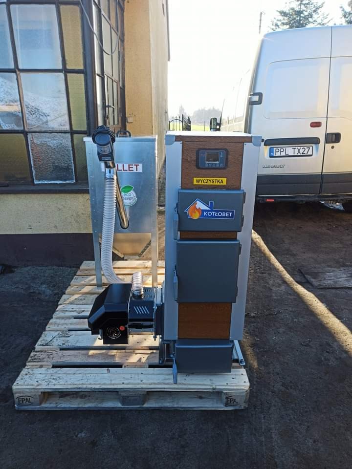 Nowoczesny piec na pellet marki Kotłobet z podajnikiem, ustawiony na palecie przed budynkiem i samochodem dostawczym.
