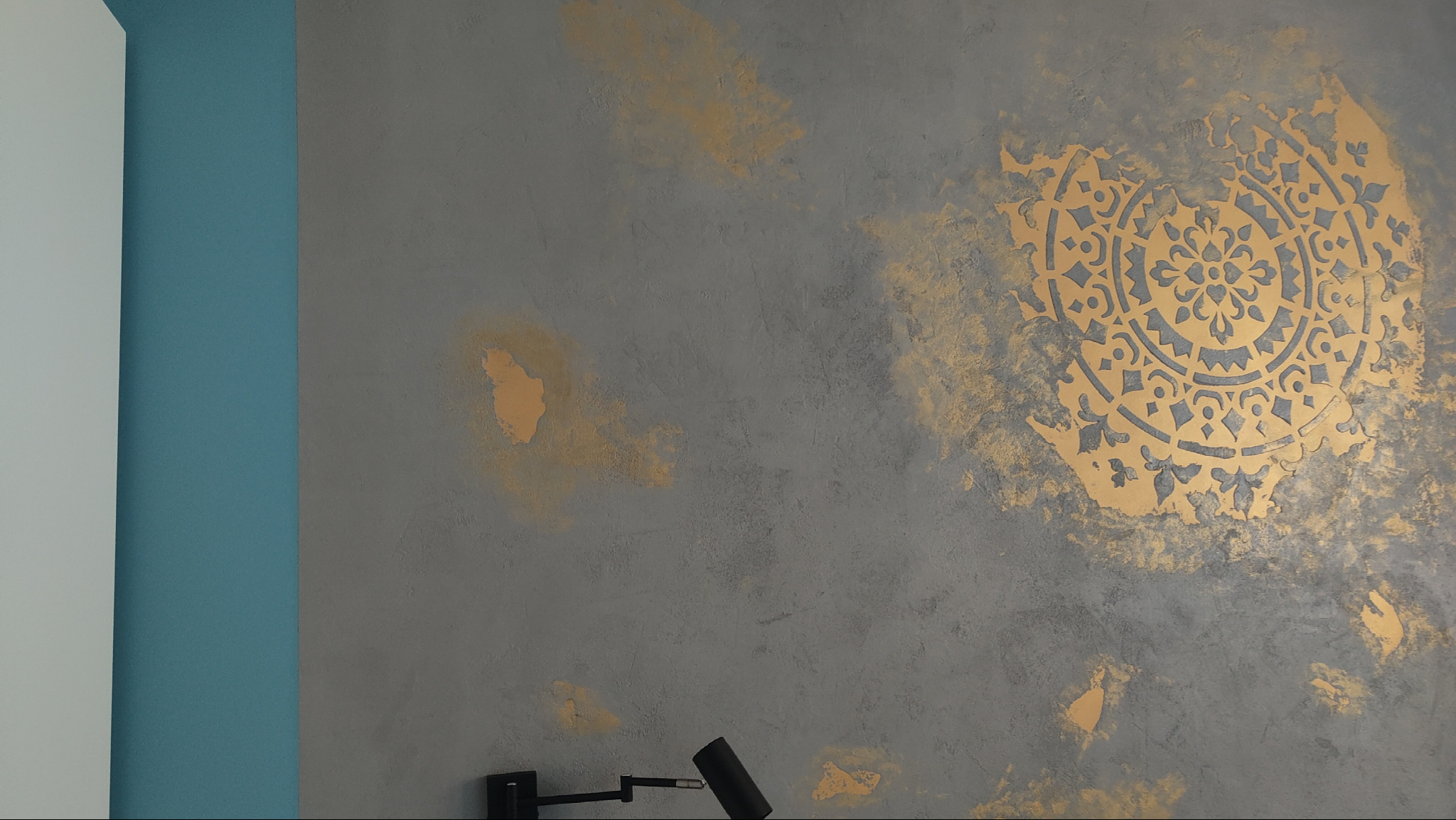 Szara ściana z dekoracyjnym, złotym ornamentem mandali, uzupełniona o minimalistyczną lampę ścienną i pionowe pasy w kolorze białym i turkusowym.