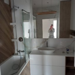 Polinbud - Wnętrze łazienki w trakcie wykańczania: biała umywalka nablatowa, bateria, lustro z odbiciem osoby robiącej zdjęcie, częściowo wykończona kabina prysznicowa, płytki imitujące marmur na ścianach...