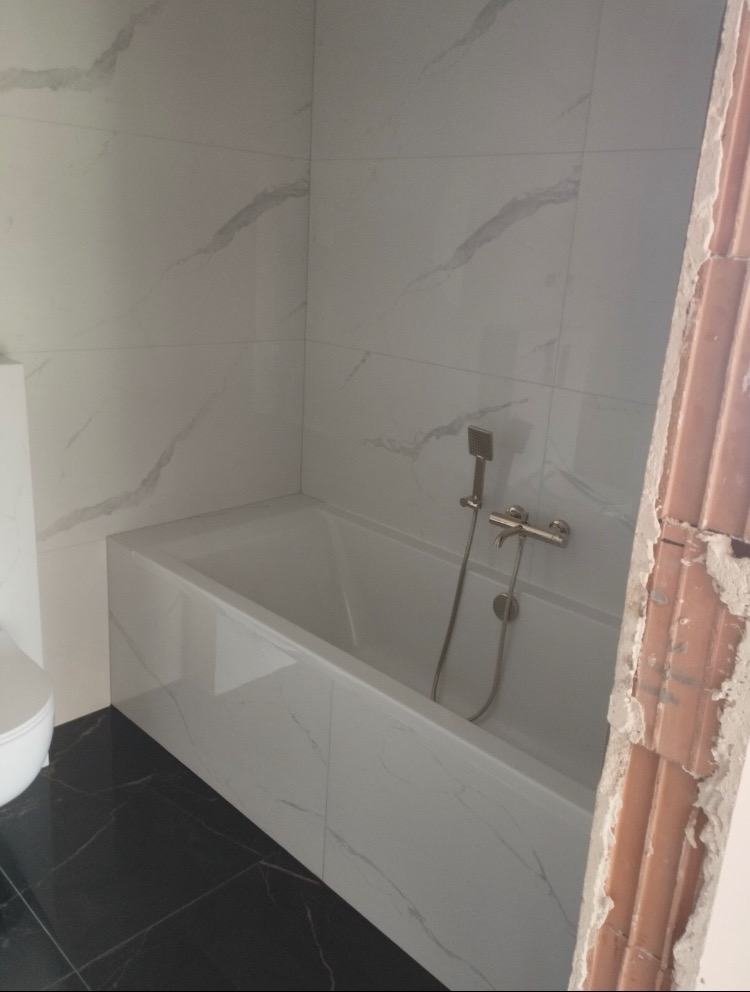 Wykończona łazienka z wanną obudowaną białymi płytkami imitującymi marmur, czarną podłogą i baterią wannową, widoczny fragment surowej ściany z cegieł.