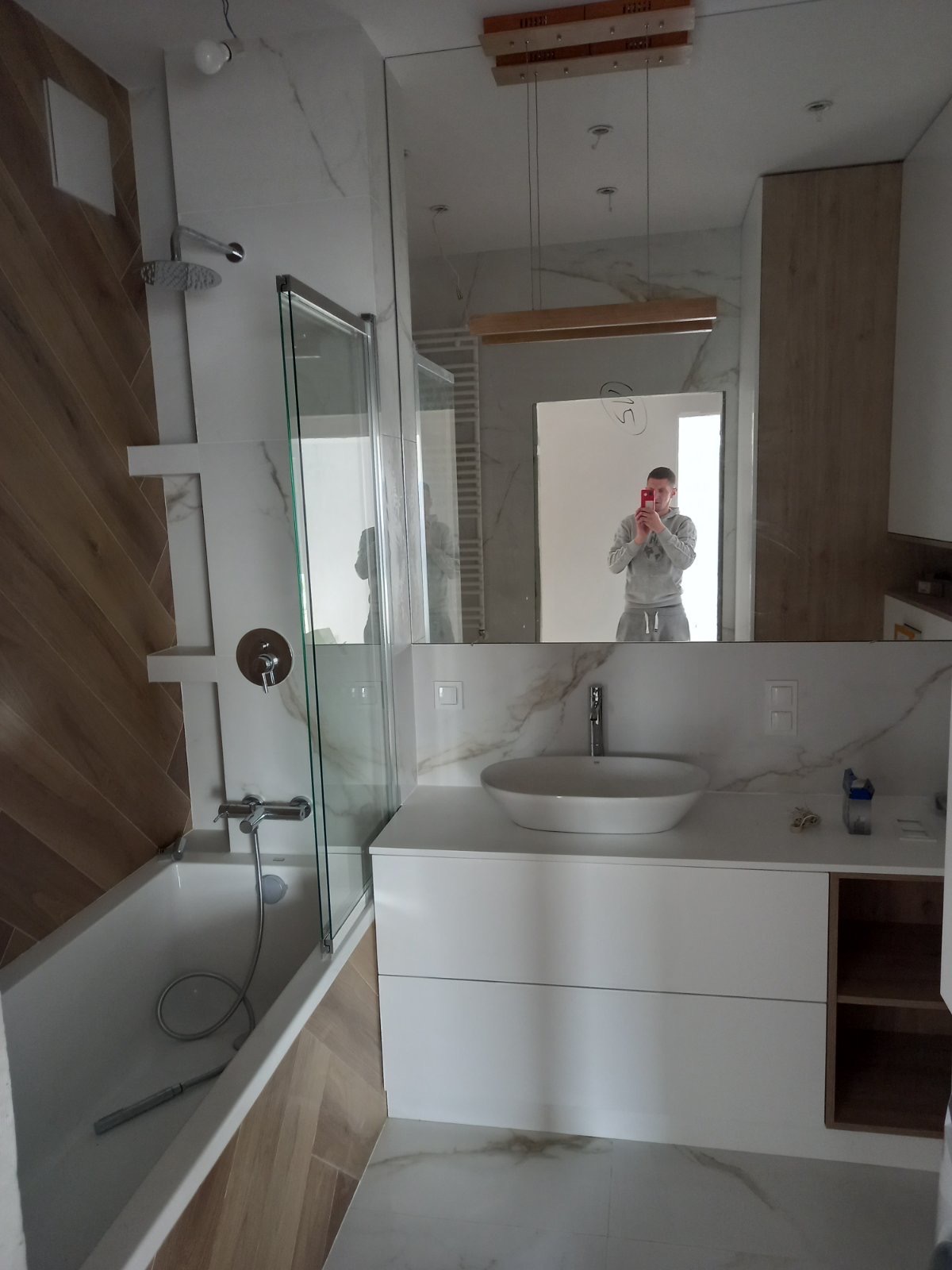 Wnętrze łazienki w trakcie wykańczania: biała umywalka nablatowa, bateria, lustro z odbiciem osoby robiącej zdjęcie, częściowo wykończona kabina prysznicowa, płytki imitujące marmur na ścianach...