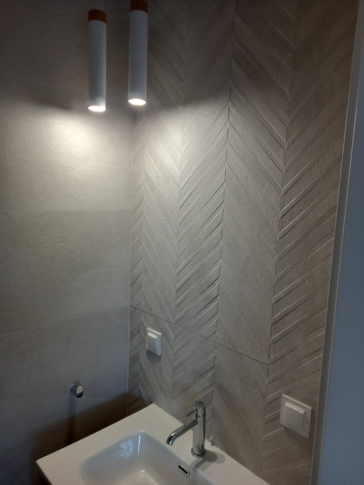 Nowoczesna łazienka w odcieniach szarości z geometrycznym wzorem jodełki na ścianie, oświetlona cylindrycznymi lampami sufitowymi, z widoczną umywalką i armaturą.